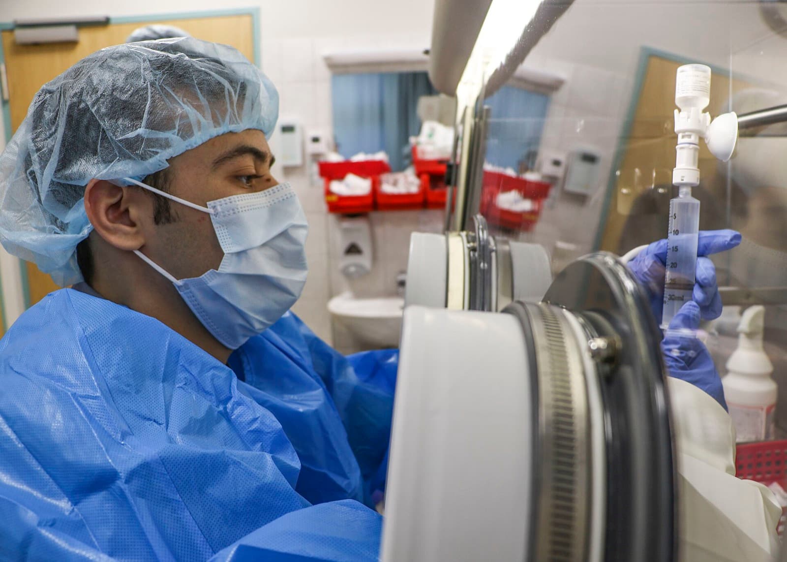 Gowned and masked pharmacy technician in sterile garb drawing a drug into a syringe inside a compounding isolator in a hospital cleanroom